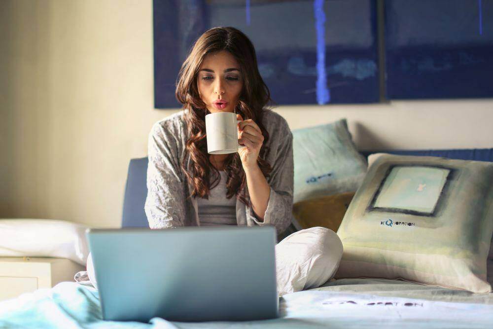 A woman on her laptop while sitting in bed, sipping on her morning coffee and enjoying her association's microlearning programs